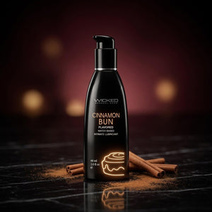 Black bottle of Wicked Cinnamon Bun flavored water-based intimate lubricant on a dark surface with cinnamon sticks.