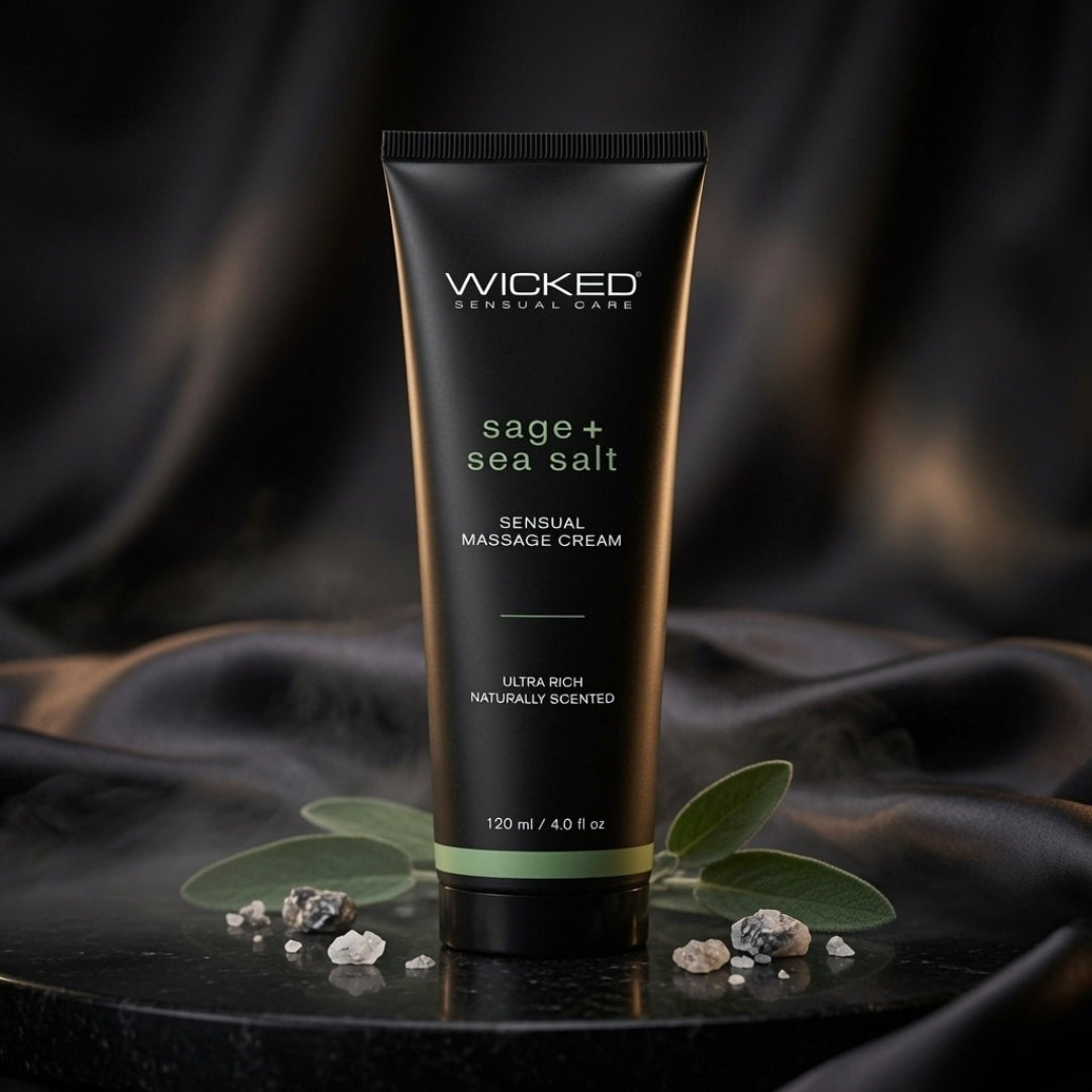 A black squeeze tube of Wicked Sensual Care 'sage + sea salt' sensual massage cream, styled on a dark reflective surface with green leaves and small stones against a draped black fabric background.