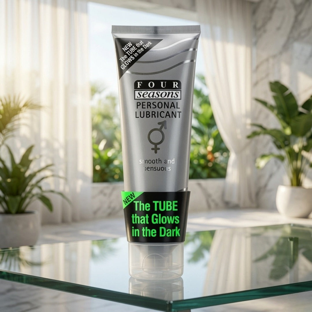 A squeeze tube of Four Seasons Personal Lubricant stands on a glass tabletop in a bright room with sheer curtains and potted plants in the background. The tube is metallic silver with black text and a black lower band featuring bright green text that reads 'The TUBE that Glows in the Dark.