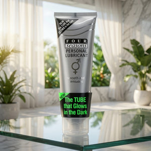 A squeeze tube of Four Seasons Personal Lubricant stands on a glass tabletop in a bright room with sheer curtains and potted plants in the background. The tube is metallic silver with black text and a black lower band featuring bright green text that reads 'The TUBE that Glows in the Dark.