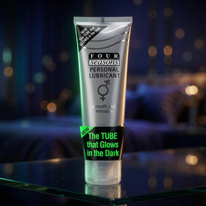 A squeeze tube of Four Seasons Personal Lubricant standing upright on a reflective glass surface against a dark, blurred background with warm bokeh lights. The tube is metallic silver with black text and a bright green 'NEW' banner near the bottom.