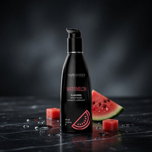 Black bottle of Wicked Watermelon flavored water-based lubricant on a dark surface with watermelon pieces.