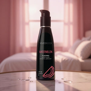 Black bottle of Wicked Watermelon flavored lubricant on a reflective surface with a blurred bedroom background.