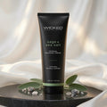 Wicked Sensual Care 'sage + sea salt' Sensual Massage Cream in a matte black squeeze tube, styled on a black marble pedestal with green leaves and sea salt crystals against a draped white fabric background.
