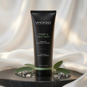 Wicked Sensual Care 'sage + sea salt' Sensual Massage Cream in a matte black squeeze tube, styled on a black marble pedestal with green leaves and sea salt crystals against a draped white fabric background.