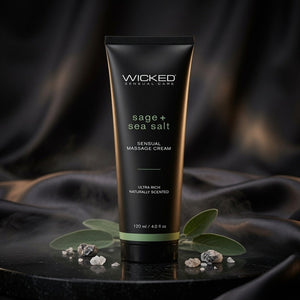 A black squeeze tube of Wicked Sensual Care 'sage + sea salt' sensual massage cream, styled on a dark reflective surface with green leaves and small stones against a draped black fabric background.