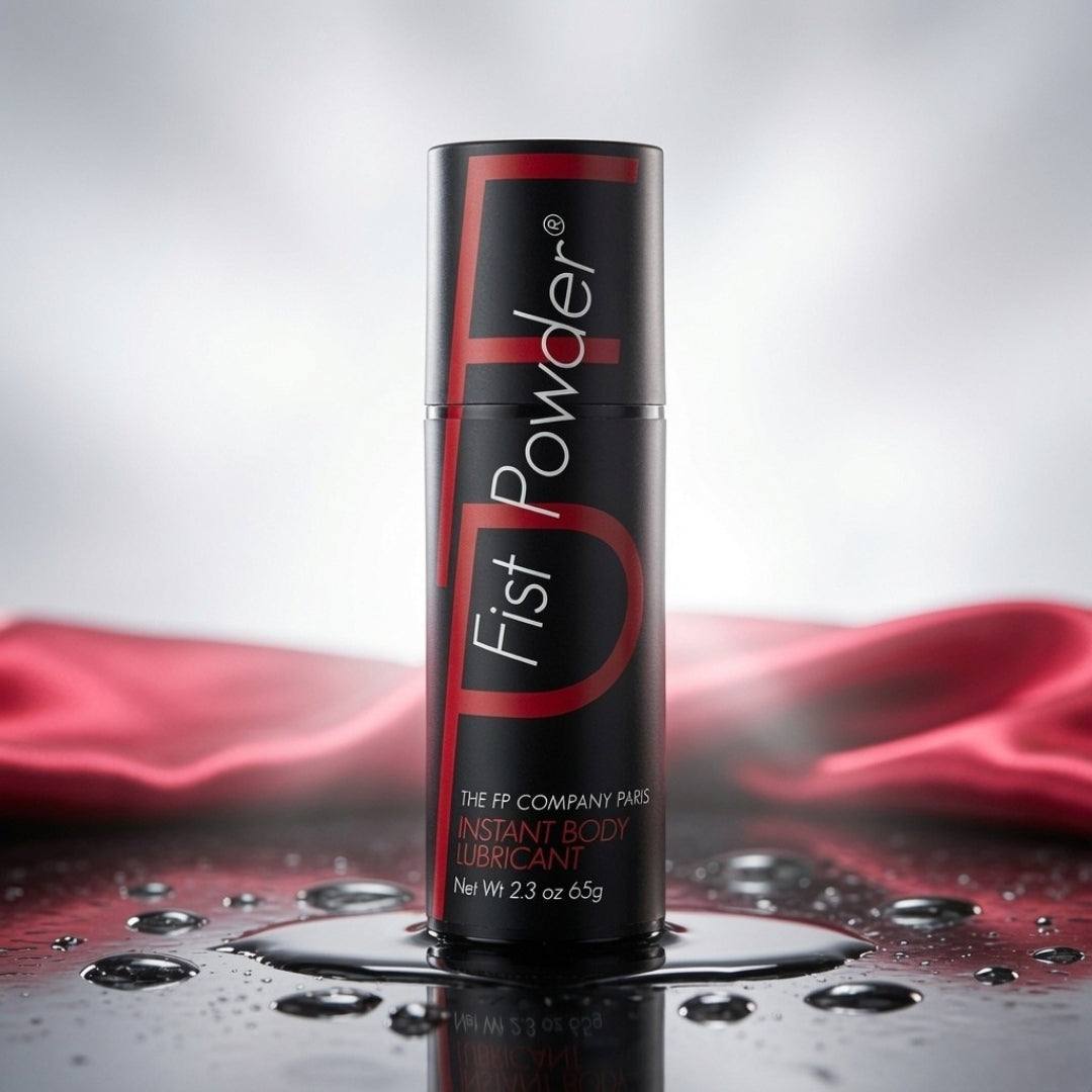 A black cylindrical bottle of Fist Powder Instant Body Lubricant with red and white lettering, standing on a glossy wet surface with water droplets and a draped red fabric in the background.